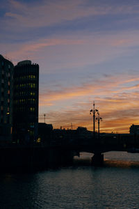 Silhouette buildings by river against sky during sunset