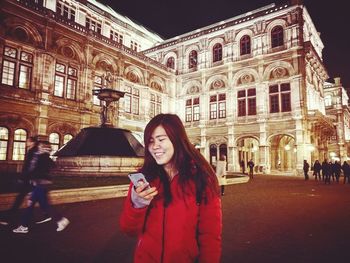 Portrait of young woman using phone while standing in front of building