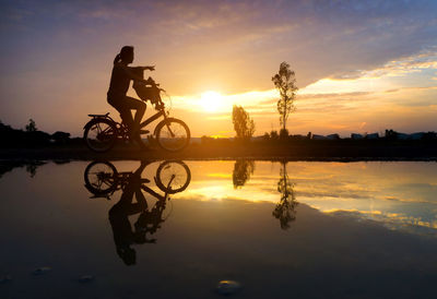 Side view of silhouette woman with son on bicycle reflecting in lake