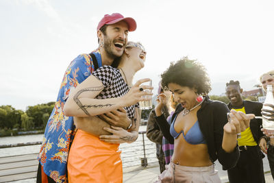 Gay man laughing while embracing non-binary friend holding drink glass in city