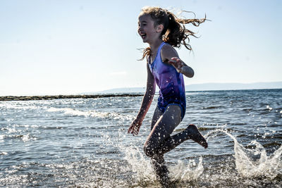 Side view of happy girl running in sea against sky during sunny day