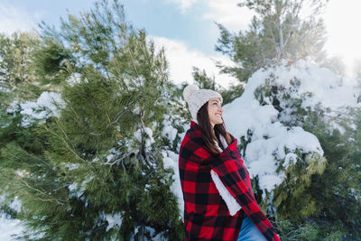 Smiling woman looking away while standing against snow covered tree