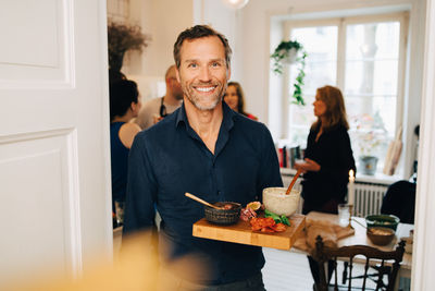 Portrait of smiling mature man holding food in serving tray while standing against friends at party