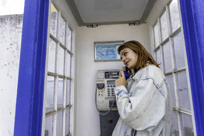 Smiling woman talking on telephone standing in telephone booth