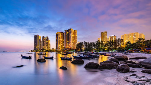 Illuminated modern buildings by sea against sky during sunset