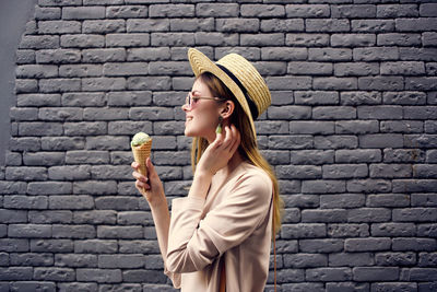 Portrait of woman holding ice cream standing against brick wall