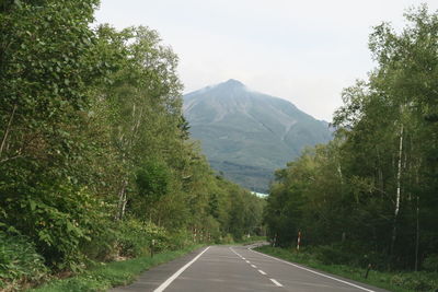 Empty road along trees and mountains against sky