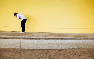 Side view of man standing on footpath against yellow wall