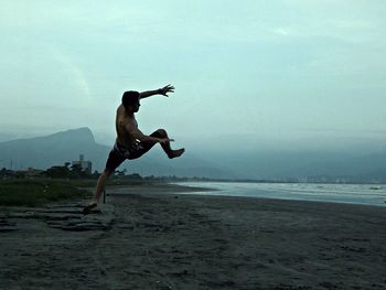 Full length of shirtless man jumping at sandy beach against sky