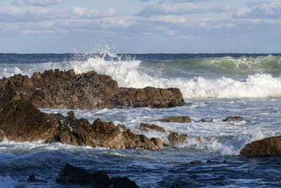 Waves splashing on rocks at shore against sky