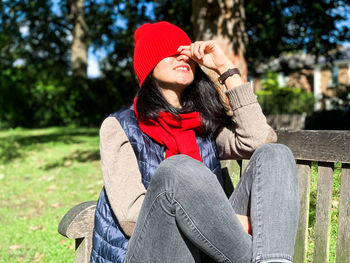 Young happy candid woman in casual autumnal clothes siting on bench in park and pulling hat down