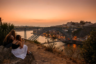 Rear view of women looking at bridge over river against orange sky during sunset