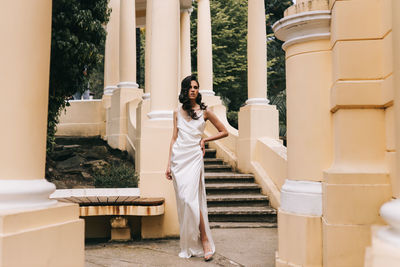 A beautiful brunette lady in an elegant wedding dress poses among the columns in the old city park