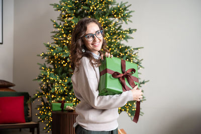 A smiling girl with glasses clutches a gift box near the christmas tree and looks away