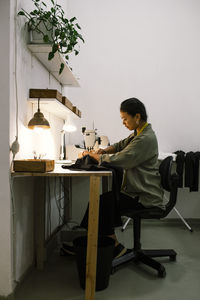 Side view of seamstress stitching clothes using sewing machine while sitting on chair at workshop