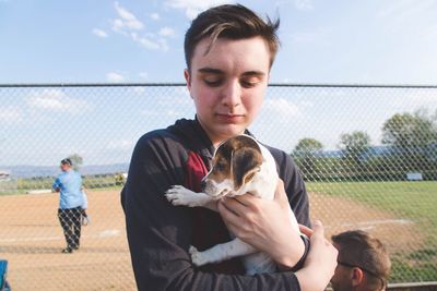 Young man carrying puppy while standing against chainlink fence