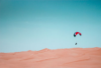 Person paragliding over sand dunes against blue sky