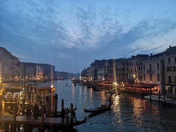 Sailboats moored on illuminated canal by buildings against sky at dusk