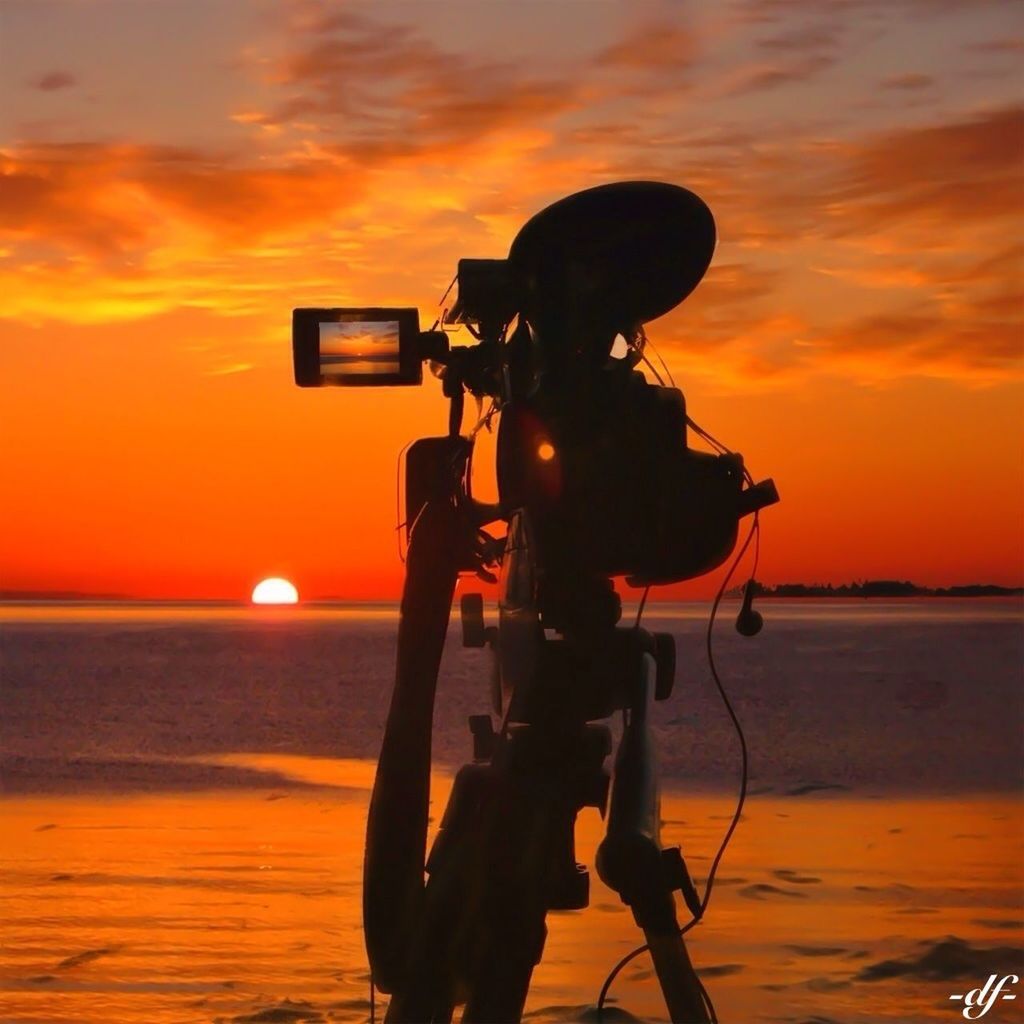 sunset, silhouette, orange color, sea, sky, lifestyles, men, sun, leisure activity, photography themes, technology, horizon over water, holding, water, standing, hobbies, camera - photographic equipment, photographing