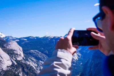 Cropped image of man photographing snowcapped mountains using phone against sky
