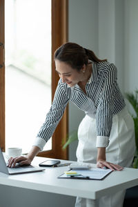 Smiling businesswoman using laptop while leaning on desk