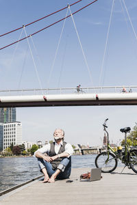 Mature man with laptop and bicycle sitting at the riverside in the city