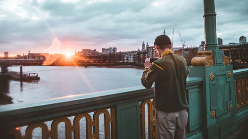Man standing on railing by river against cityscape during sunset
