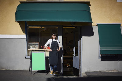 Confident female owner standing with hands on hips at entrance of store