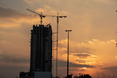 Low angle view of crane by building against sky during sunset