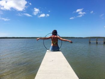 Rear view of young woman with plastic hoop sitting on pier over lake against sky during sunny day