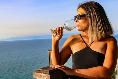 Portrait of young woman drinking glass while standing on beach