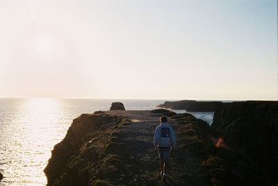 Rear view of man walking on cliff at sea against clear sky during sunset