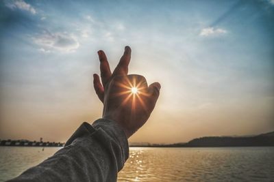 Sunbeam streaming through cropped hand by sea against sky