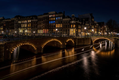 Illuminated bridge over river by buildings against sky at night