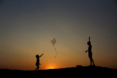 Silhouette children flying kite against clear sky during sunset