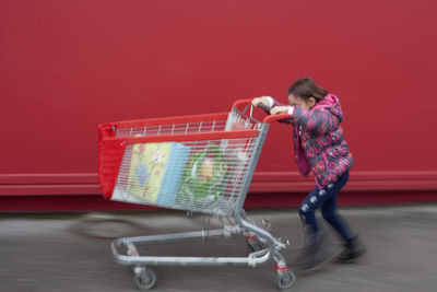 Playful girl pushing shopping cart against red wall