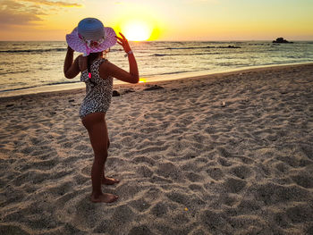 Side view of girl wearing hat standing at beach during sunset