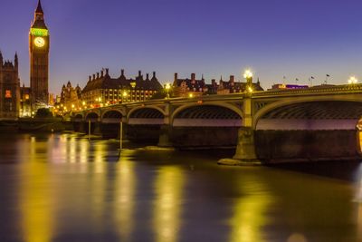 Illuminated bridge over river by buildings against sky at night