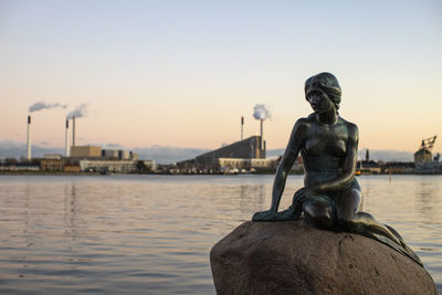 Statue by river against sky during sunset