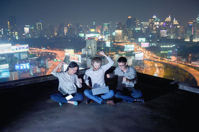 Happy colleagues sitting on building terrace against illuminated city at night