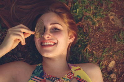 High angle portrait of smiling woman covering eye with leaf while lying at park