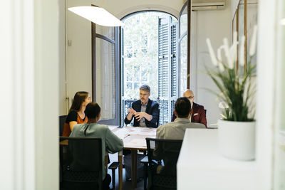 Businessman gesturing while discussing with coworkers at table in office seen through doorway