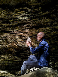 Side view of man holding stone while sitting in cave