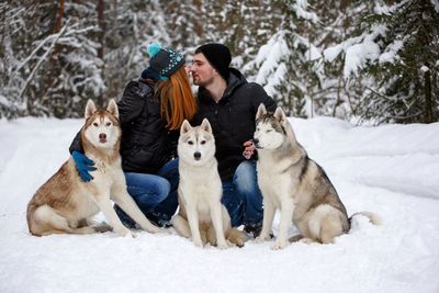 Young couple kissing on snowy field with siberian huskies against trees