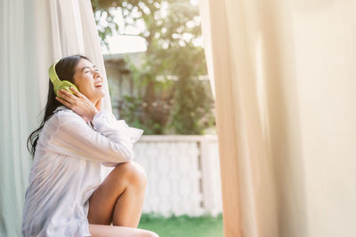 Side view of woman listening music while sitting by window at home