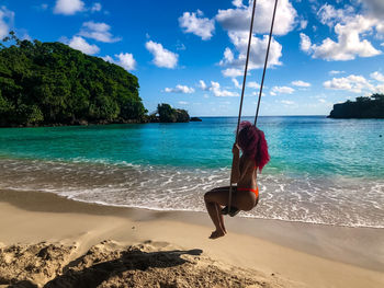 Full length of woman sitting on rope swing at beach against sky