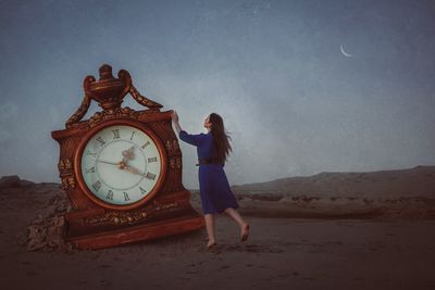 Full length of woman touching clock on sand at beach against sky