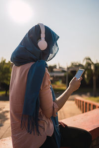 Side view of arab female in headscarf and headphones browsing mobile phone and listening to music while enjoying sunny day in city