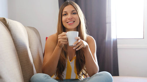 Portrait of young woman holding coffee cup while sitting on sofa at home