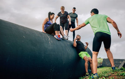 Friends assisting each other by pipe against sky